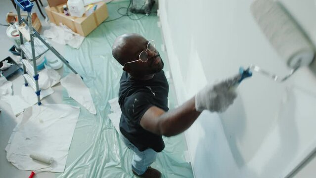 Black man standing on the floor covered with cellophane and painting a wall with roller during DIY apartment renovation. High angle shot