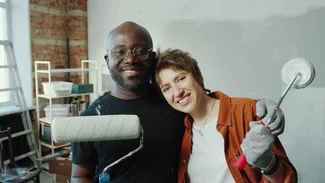 Happy diverse couple holding paint rollers, embracing and smiling at camera in living room under renovation. Zoom shot, video portrait