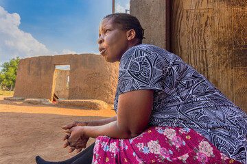 village african woman with braids in front of her home, sited on the veranda, ruins of mud house in...