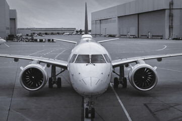 Front view of a modern twinjet airliner sitting on the concrete tarmac with lane markings in a large airport. Black and white - obrazy, fototapety, plakaty