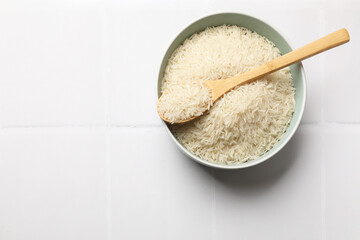 Raw basmati rice in bowl and spoon on white tiled table, top view. Space for text