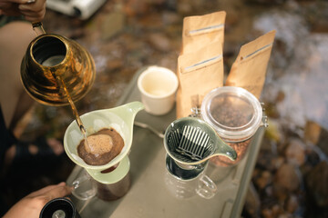 A man is pouring coffee into a cup from a gold pot. The scene is set in a natural environment, with a stream nearby. The man is enjoying the moment, as he pours the coffee into the cup