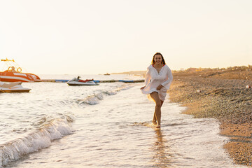 A smiling young woman running along a sandy shoreline with water splashing around her feet. The evening sun bathes the scene in a warm glow with several boats floating in the distant background