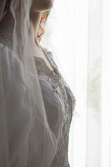 Close up of the bodice of a vintage wedding dress against a window with a lace curtain. The dress was photographed on a mannequin with the neck and chin of the woman created using AI 