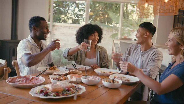 Group of multi-racial friends sitting around table enjoying meal at dinner party making cheers with water- shot in slow motion