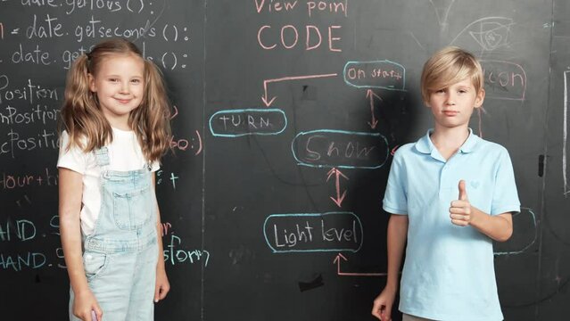 Caucasian girl writing blackboard with engineering prompt or coding, programing system written in STEM technology classroom and turnaround to looking at camera while boy showing thumb. Erudition.