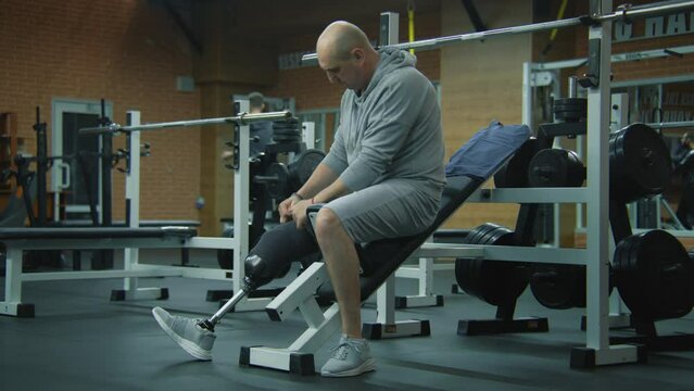 Motivated athlete with prosthetic leg corrects prosthesis and starts to train with empty barbell in modern gym. Fit man with physical disability does exercises using professional sports equipment.