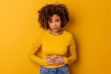 a worried woman in a yellow shirt touching her stomach on yellow studio background - obrazy, fototapety, plakaty
