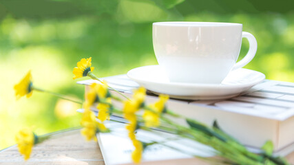 White steaming cup of hot coffee owith yellow flowers and green nature blurred background.