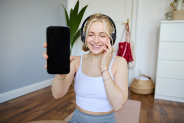 Portrait of smiling, beautiful young woman, showing her smartphone screen, wearing headphones, looking satisfied with sound quality - obrazy, fototapety, plakaty