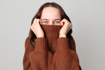 Shy funny brown haired woman wearing brown jumper standing isolated over light gray background hiding half of her face with collar of sweater looking at camera with happy eyes