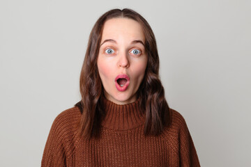 Shocked brown haired woman wearing brown jumper standing isolated over light gray background looking at camera with big eyes and open mouth say wow OMG