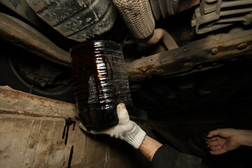 Black engine oil flows into a canister held by a mechanic under the bottom of the car. A mechanic...