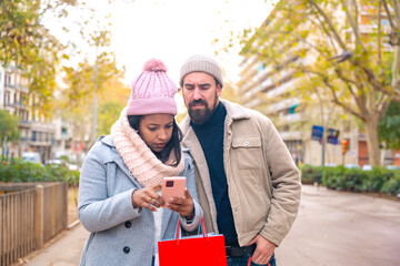 Cute multiethnic couple of man and woman standing holding shopping bags, browsing smartphone apps shopping online