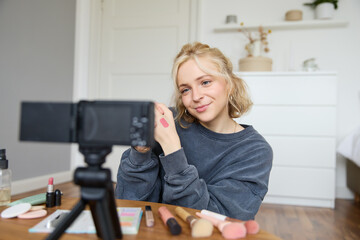 Image of young stylish woman, blogger recording a beauty lifestyle video of her picking best lipstick, showing lip balm swatches on her skin, sitting in front of digital camera in empty room
