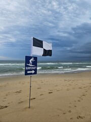 Cloudy Beach Day: black and white flag marks designated surfcraft area, waves gently crashing as footprints scatter across the beach - obrazy, fototapety, plakaty