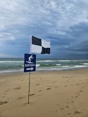 Fototapeten Cloudy Beach Day: black and white flag marks designated surfcraft area, waves gently crashing as footprints scatter across the beach © Jam-motion