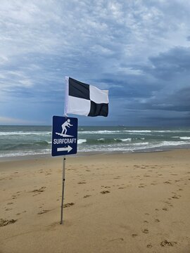 Fototapeta Cloudy Beach Day: black and white flag marks designated surfcraft area, waves gently crashing as footprints scatter across the beach