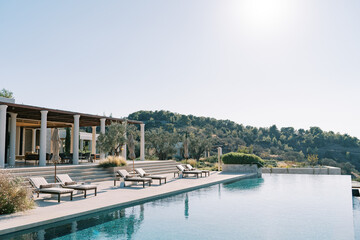 Fototapeta na wymiar Sun loungers with folded sun umbrellas line the pillared terrace next to the long swimming pool. Hotel Amanzoe. Peloponnese, Greece
