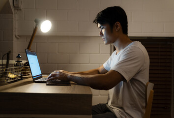 Stressed young Asian man on a desk looking at a laptop computer in darkness late at night, working,...