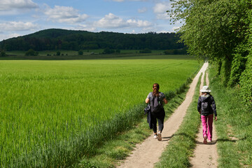 Zwei Frauen mit Rucksäcken wandern auf einem Feldweg durch frühlingshafte Landschaft