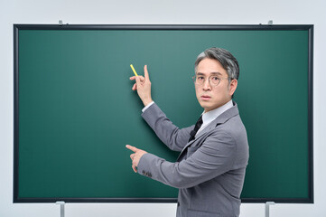 A middle-aged male teacher in a suit and glasses is giving an Internet lecture with a confident expression and pose with chalk in front of a green blackboard.
