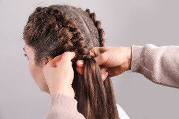 Professional stylist braiding woman's hair on grey background, closeup - obrazy, fototapety, plakaty