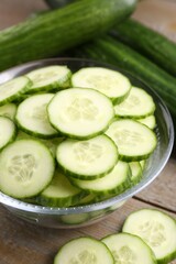 Cut cucumber in glass bowl and fresh vegetables on wooden table, closeup