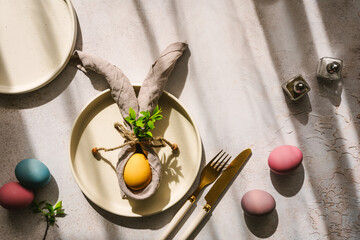 Ein gelbes Osterei mit Hasenohren aus einer Stoffserviette auf einem Teller im Sonnenlicht. Draufsicht, Ostern Hintergrund. - obrazy, fototapety, plakaty