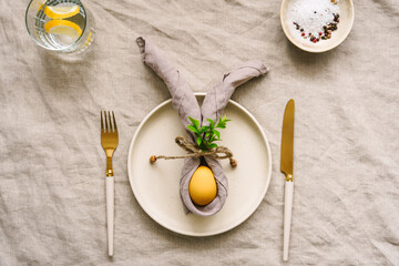 Ein gelbes Osterei mit Hasenohren aus einer Stoffserviette auf einem Teller. Messer und Gabel,...