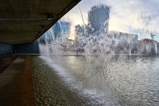 View from behind of the water falling from a waterfall of some buildings that are seen through the water of a park without people