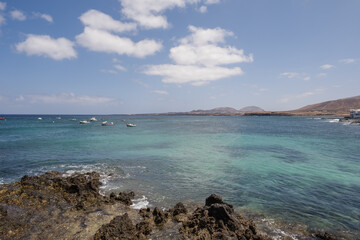 Seascape. Rocks in the foreground. Group of boats anchored nearby. Mountains in the background. Turquoise Atlantic Ocean. Big white clouds. Village of Arrieta. Lanzarote, Canary Islands, Spain