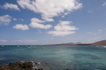 Seascape. Rocks in the foreground. Group of boats anchored nearby. Mountains in the background. Turquoise Atlantic Ocean. Big white clouds. Village of Arrieta. Lanzarote, Canary Islands, Spain