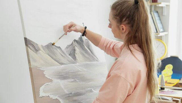 Young woman painter drawing picture on canvas using acrylic paints. Female artist works in studio. Landscape, sea, mountains. Drawing concept. Hobby. Canvas stands on easel. 4K, UHD