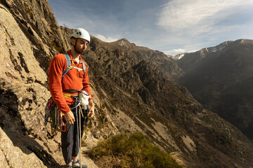 A man in an orange shirt is standing on a rocky mountain. He is wearing a helmet and has a backpack on. The mountain is covered in snow and the sky is clear