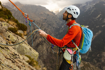 A man in a red jacket is climbing a mountain with a blue backpack. He is wearing a helmet and has a...