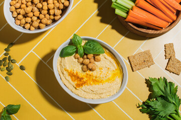 Delicious chickpea hummus in a plate on a bright yellow background with fresh vegetables. 