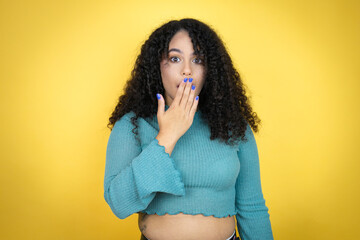 African american woman wearing casual sweater over yellow background covering mouth with hand,...