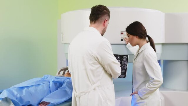 Doctors discuss and consult with each other against the backdrop of an MRI scanner and a young female patient in a modern clinic. Concept of professional communication and medical technologies.