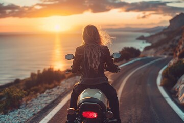 Rear view of a young long haired woman without a helmet riding a motorcycle on an asphalt road near the sea under sunset lights - obrazy, fototapety, plakaty