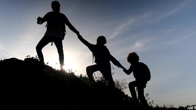 Mother woman helps her two children climb the mountain holding hands in the shape of a chain, sunset silhouette.