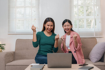 Two young women joyfully celebrate a successful moment together in front of a laptop in a cozy home...
