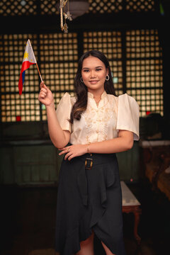 Portrait of a confident young woman holding a Philippine flag, dressed in elegant traditional Filipino attire.