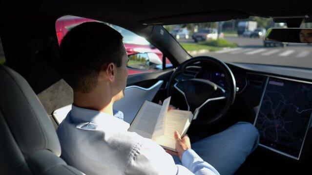 Male businessperson reading book during riding on electrical vehicle with autopilot at urban road. Successful businessman improving his knowledge while riding an autonomous self driving electric car