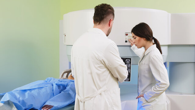 Doctors discuss and consult with each other against the backdrop of an MRI scanner and a young female patient in a modern clinic. Concept of professional communication and medical technologies.