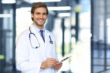 Handsome friendly young doctor on hospital corridor looking at camera, smiling. - obrazy, fototapety, plakaty