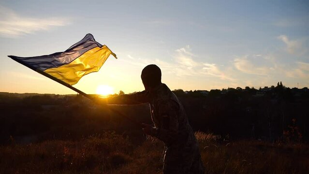 Young man in military uniform waving flag of Ukraine against sunset at background. Male ukrainian army soldier lifted national banner at countryside. Victory against russian aggression. End of war