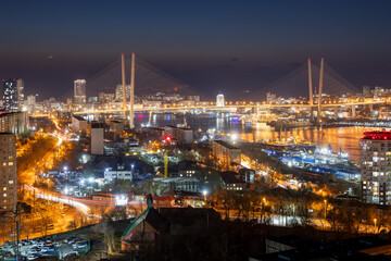 Beautiful night city landscape. Top view of streets and buildings. In the distance is a large cable-stayed Golden Bridge over the Golden Horn Bay. Vladivostok city, Primorsky Krai, Russian Far East.