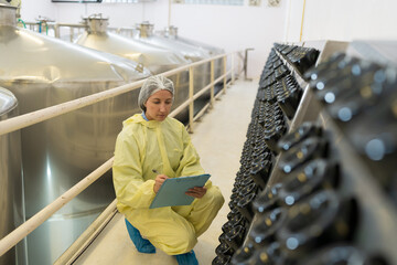 Female factory worker inspecting quality of wine bottles on shelf during manufacturing in process...