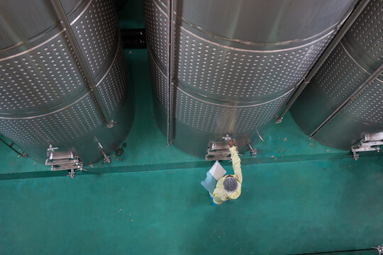 Winemaker working in wine factory. Top view of factory worker inspecting quality of stainless tank of wine fermented during manufacturing in process. Wine manufacturing industry concept